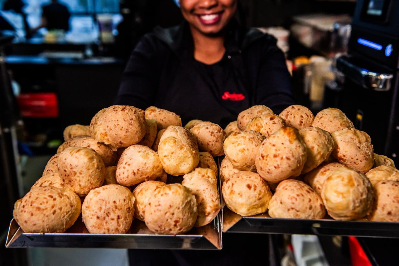 Atendente da Suíça sorrindo, segurando duas bandejas cheias de pão de queijo recém-assado.